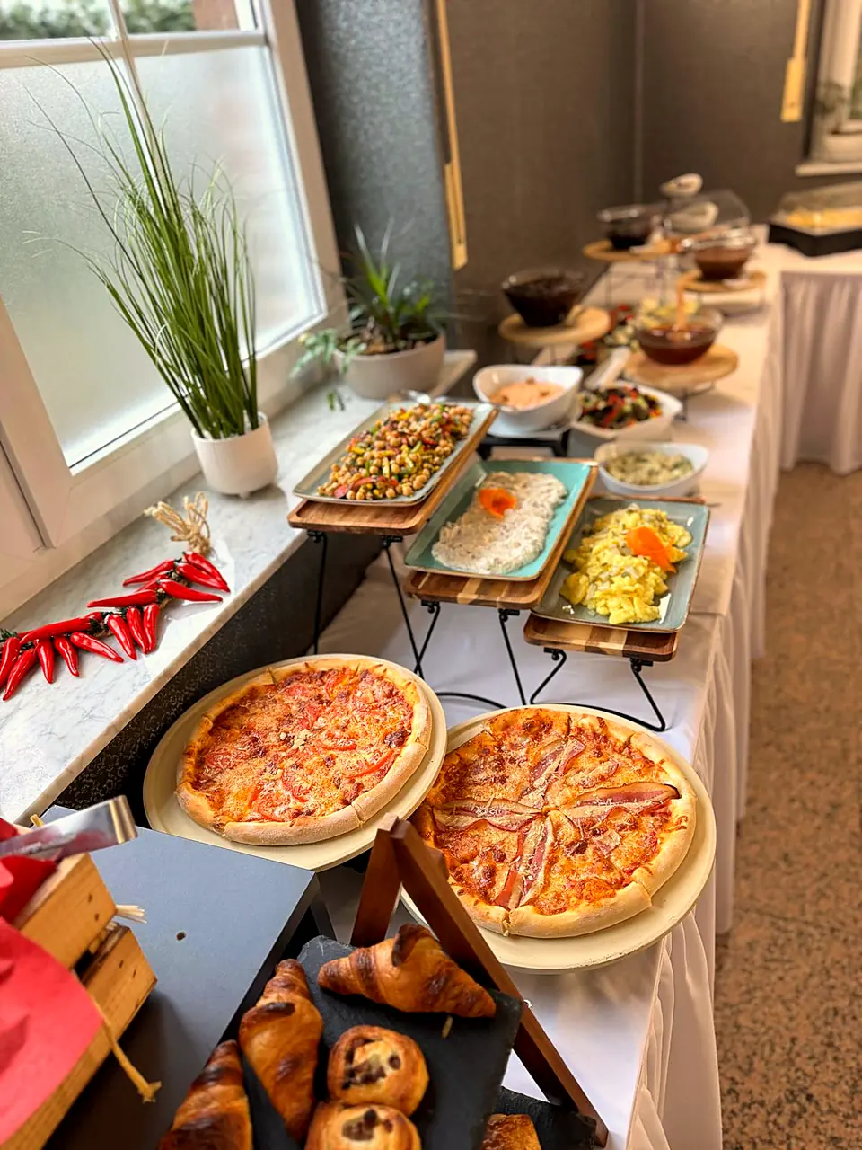 Buffet mit Pizza, Croissants, Salaten und Dips am Fenster